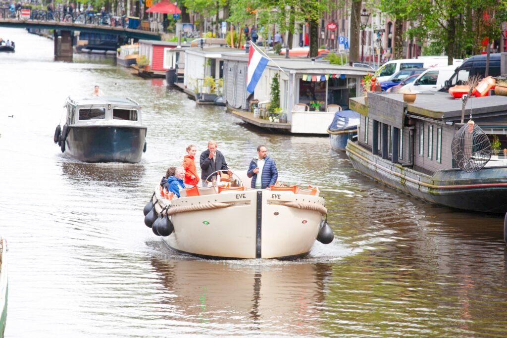 Amsterdam canal with houseboats and tourist boat cruising under green trees.