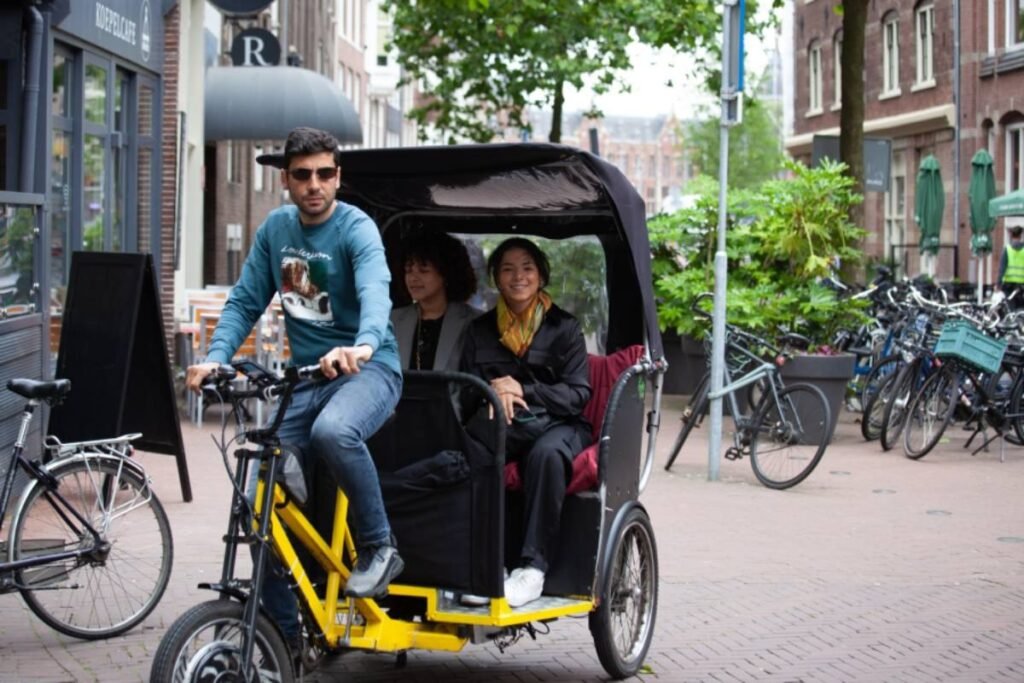 Driver pedaling bright yellow Amsterdam bike taxi pedicab with smiling couple enjoying private tour on city street.