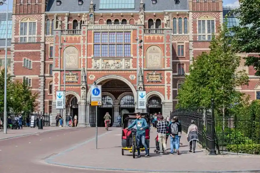 Ornate Rijksmuseum facade with tourists and bike taxi in background under trees in Amsterdam.
