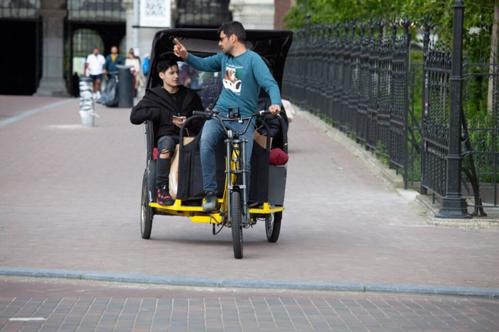 Passengers riding yellow bike taxi pedicab past Anne Frank House entrance and sign in central Amsterdam.