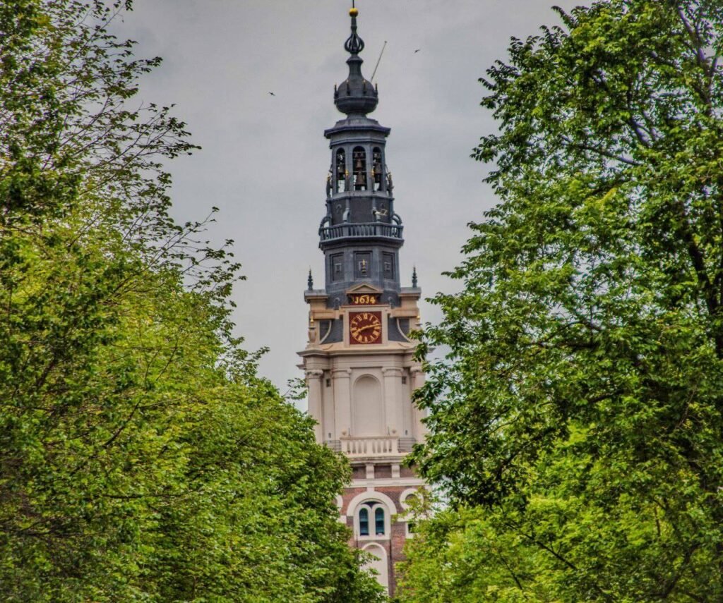 Iconic Westerkerk tower rising above green trees on a cloudy day in Amsterdam historic center.